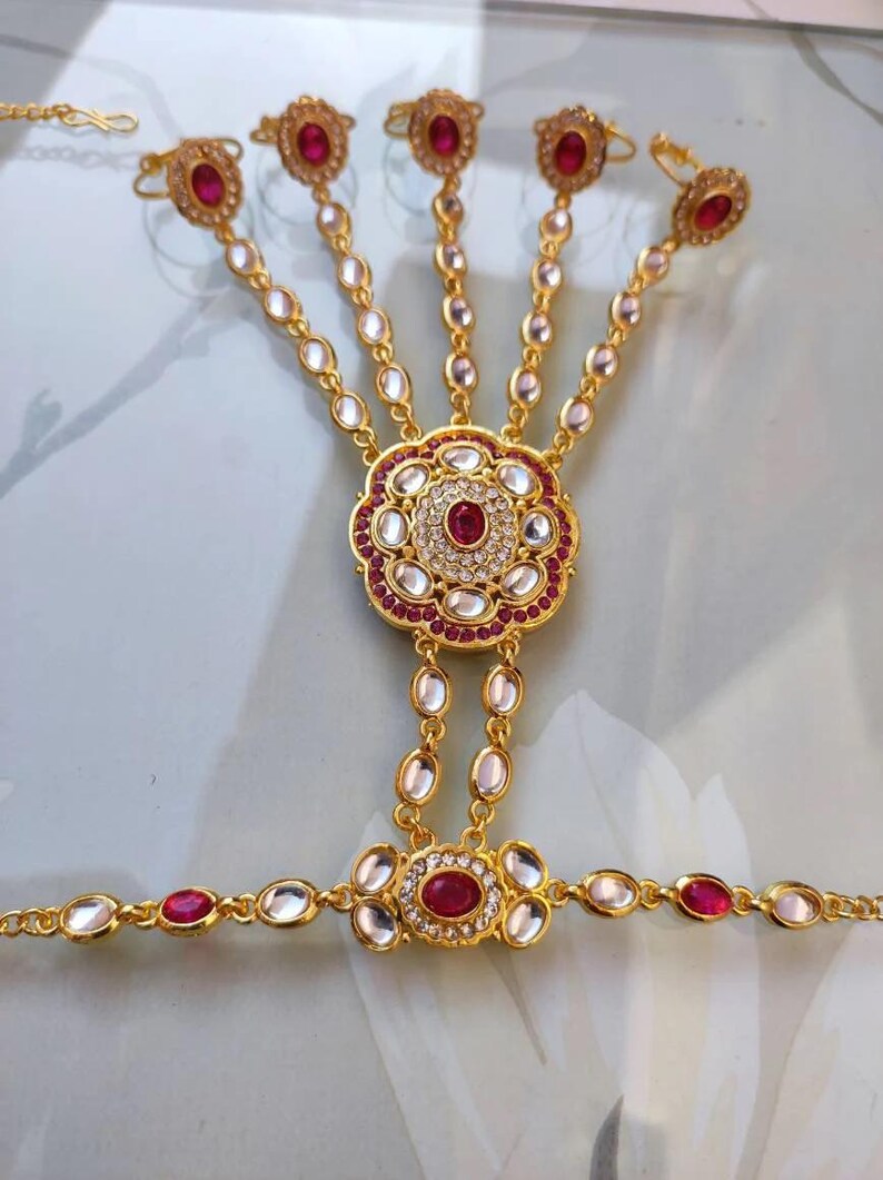 Pu&ograve; includere: Gioiello per mano dorato con un motivo floreale centrale con una grande gemma rossa circondata da pietre bianche e rosse pi&ugrave; piccole. Il pezzo si collega a cinque anelli per le dita tramite delicate catene, ognuno con una gemma rossa.