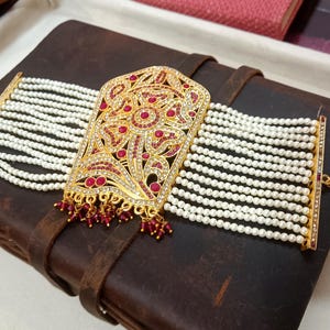May include: A gold-toned bracelet with a central rectangular design, adorned with red gemstones and clear crystals. The bracelet features multiple strands of white pearls and is set against a dark brown leather-bound book.