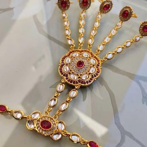 Pu&ograve; includere: Gioiello per mano dorato con pietre preziose rosse e chiare. Il pezzo presenta un motivo floreale centrale e anelli per le dita collegati da delicate catene. Un accessorio tradizionale.