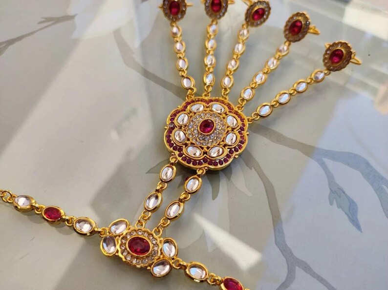 Pu&ograve; includere: Gioiello per mano dorato con pietre preziose rosse e chiare. Il pezzo presenta un motivo floreale centrale e anelli per le dita collegati da delicate catene. Un accessorio tradizionale.