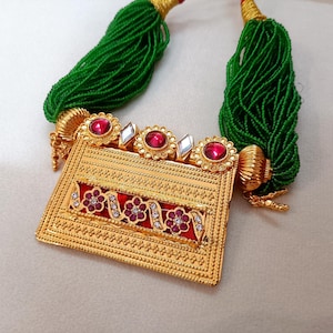 Può includere: Una collana dorata con un pendente rettangolare e fili di perline verdi. Il pendente presenta pietre preziose rosse e motivi floreali. La collana ha una superficie dettagliata e strutturata, probabilmente in metallo.