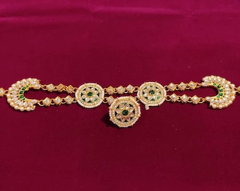 May include: A gold-coloured jewellery item with a central design and two crescent-shaped ends. It features green and white detailing, small clear stones, and a pearl-like border. The background is a deep red.