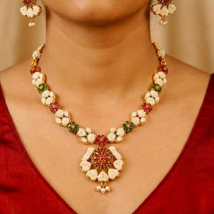 Könnte beinhalten: Ein goldfarbenes Halsketten- und Ohrring-Set. Die Halskette hat ein Blumenmuster mit weißen, roten und grünen Akzenten. Das Model trägt ein rotes Oberteil. Der Schmuck ist wahrscheinlich für besondere Anlässe.