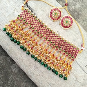 May include: A gold-toned necklace and earring set. The necklace features a geometric pattern with red and green accents, small white pearls, and dangling green beads. The earrings are circular with red and green details.