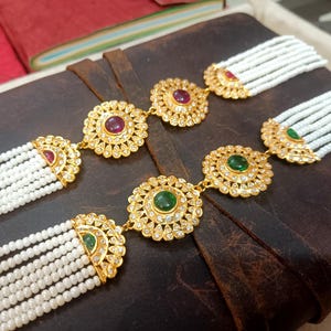 Może przedstawiać: Zbliżenie dwóch złotych naszyjników z białymi sznurami koralików. Każdy naszyjnik posiada trzy okrągłe, złote wisiorki z centralnym kamieniem szlachetnym, otoczone mniejszymi, przezroczystymi kamieniami. Wisiorki połączone są złotymi ogniwami.