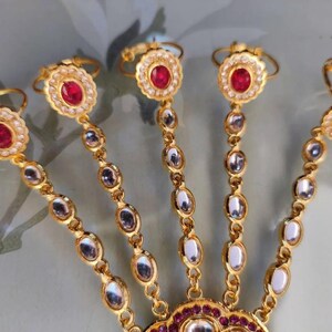 Pu&ograve; includere: Gioiello per mano dorato con pietre preziose rosse e trasparenti. Il design include anelli per ogni dito collegati da catene di pietre ovali. Il pezzo centrale presenta un design grande e ornato con altre pietre rosse.