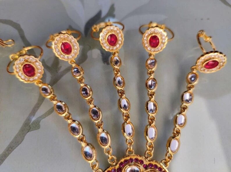 Pu&ograve; includere: Gioiello per mano dorato con pietre preziose rosse e trasparenti. Il design include anelli per ogni dito collegati da catene di pietre ovali. Il pezzo centrale presenta un design grande e ornato con altre pietre rosse.