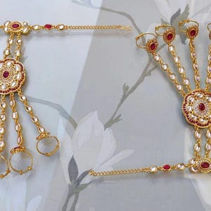 Pu&ograve; includere: Gioielli per le mani color oro con accenti di pietre preziose rosse e chiare. Il design presenta un motivo floreale centrale e catene che collegano agli anelli. I gioielli sono probabilmente per occasioni speciali.