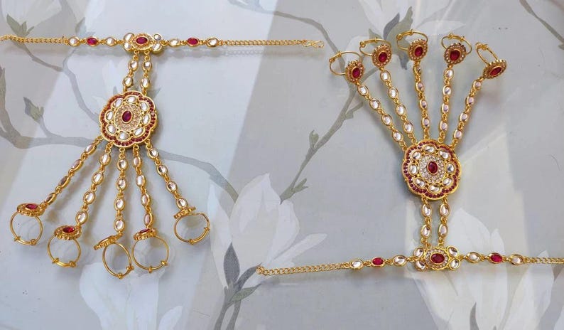 Pu&ograve; includere: Gioielli per le mani color oro con accenti di pietre preziose rosse e chiare. Il design presenta un motivo floreale centrale e catene che collegano agli anelli. I gioielli sono probabilmente per occasioni speciali.