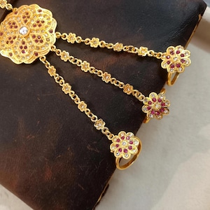 May include: A gold-toned hand chain with a floral design. The chain features a large, ornate flower pendant with a central clear stone and smaller red stones. Three delicate chains extend to rings, each with a flower-shaped ring adorned with red and clear stones.