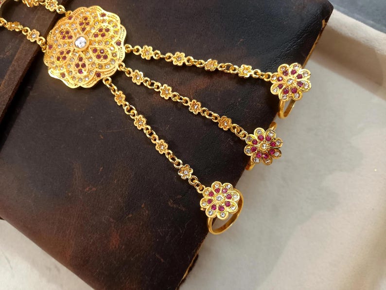 May include: A gold-toned hand chain with a floral design. The chain features a large, ornate flower pendant with a central clear stone and smaller red stones. Three delicate chains extend to rings, each with a flower-shaped ring adorned with red and clear stones.