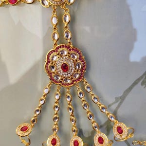 Pu&ograve; includere: Gioiello per mano dorato con pietre preziose rosse e trasparenti. Il design presenta un motivo floreale centrale e anelli per le dita collegati da delicate catene. Il pezzo &egrave; ornato e probabilmente destinato a occasioni speciali.