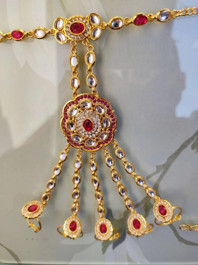 Pu&ograve; includere: Gioiello per mano dorato con pietre preziose rosse e trasparenti. Il design presenta un motivo floreale centrale e anelli per le dita collegati da delicate catene. Il pezzo &egrave; ornato e probabilmente destinato a occasioni speciali.