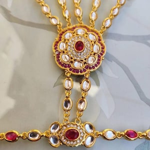 Pu&ograve; includere: Una catena per la mano color oro con un motivo floreale centrale con una pietra centrale rosso rubino e pietre preziose chiare e rosse circostanti. La catena si estende ad anelli per le dita e un braccialetto da polso, tutti adornati con pietre preziose simili.