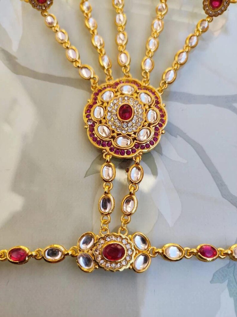 Pu&ograve; includere: Una catena per la mano color oro con un motivo floreale centrale con una pietra centrale rosso rubino e pietre preziose chiare e rosse circostanti. La catena si estende ad anelli per le dita e un braccialetto da polso, tutti adornati con pietre preziose simili.