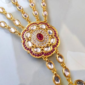 Pu&ograve; includere: Gioiello per mano dorato con un design floreale centrale, una pietra centrale color rubino e dettagli chiari e color rubino. Il pezzo ha pi&ugrave; catene con pietre ovali trasparenti.