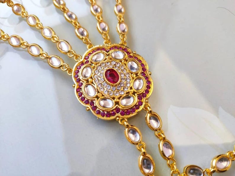 Pu&ograve; includere: Gioiello per mano dorato con un design floreale centrale, una pietra centrale color rubino e dettagli chiari e color rubino. Il pezzo ha pi&ugrave; catene con pietre ovali trasparenti.