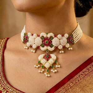 Puede incluir: Primer plano de un collar gargantilla de perlas blancas con un diseño floral. El collar presenta detalles en rojo y verde y un colgante en forma de corazón. La modelo lleva un sari rojo y dorado.