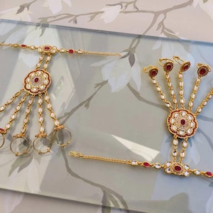 Pu&ograve; includere: Due gioielli per le mani color oro. Ognuno presenta un motivo floreale centrale con accenti rossi e bianchi, collegato da catene ad anelli per le dita. I gioielli sono adornati con pietre preziose rosse e perle bianche.