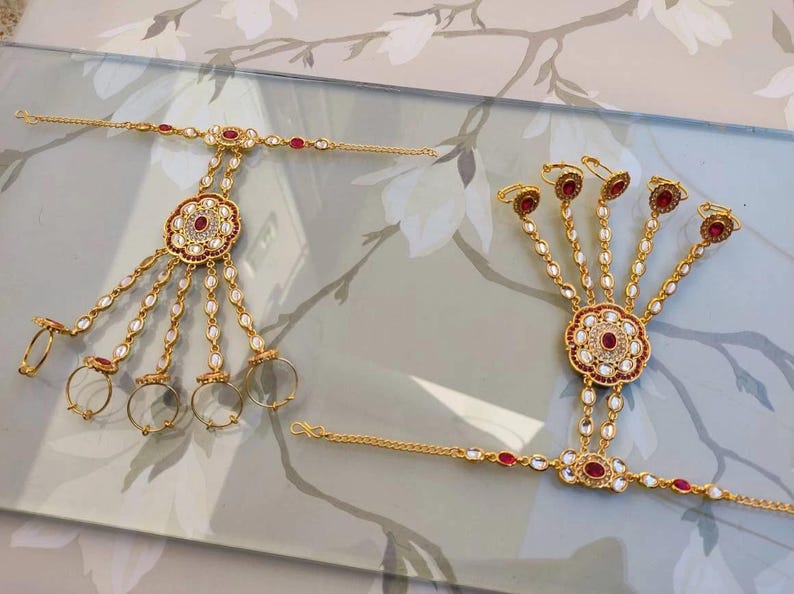 Pu&ograve; includere: Due gioielli per le mani color oro. Ognuno presenta un motivo floreale centrale con accenti rossi e bianchi, collegato da catene ad anelli per le dita. I gioielli sono adornati con pietre preziose rosse e perle bianche.