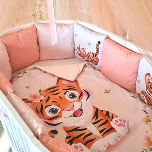 Puede incluir: Un juego de cuna para bebé con diseño de tigre. El juego incluye una sábana bajera, protectores y almohadas decorativas en tonos melocotón y blanco. Una capota transparente añade un toque suave y aireado.
