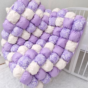 Peut inclure: Une couverture de bébé faite à la main, gonflante, dans une palette de couleurs lilas et crème. La couverture présente un motif matelassé avec des carrés de tissu lilas unis et crème à motifs alternés. La couverture est exposée sur un lit bébé blanc.