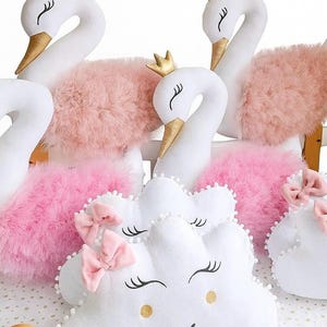 May include: Soft white swan toys with gold crowns and beaks, and pink fluffy details. Cloud-shaped cushions with pink bows and smiling faces are also shown. These items are designed for a nursery or children's room.