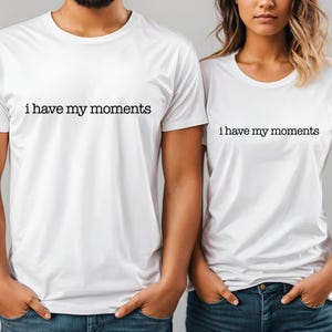 May include: Two white t-shirts with the phrase "i have my moments" printed in black. The shirts are being worn by two people, and they are both wearing blue jeans. The shirts have short sleeves and a crew neck.