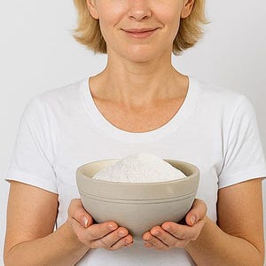 May include: A light gray ceramic bowl filled with a large quantity of white powder. The bowl is held by a person wearing a white t-shirt. The background is a plain white.