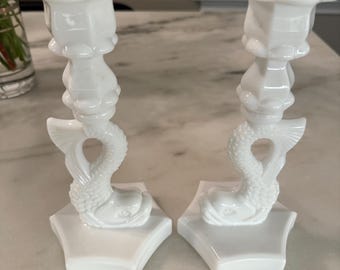 May include: A pair of white ceramic candle holders. Each has a dolphin design and a flower-shaped top. The candle holders are set on a white, hexagonal base.