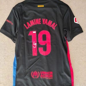 May include: Black athletic jersey with the name "LAMINE YAMAL" and the number "19" in red. The jersey has blue and red side accents, and the UNHCR logo is visible. The sleeve has the word "LIGHT" printed on it.