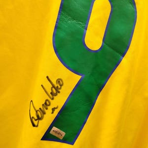 K&ouml;nnte beinhalten: Gelbes Trikot mit einer gro&szlig;en gr&uuml;nen Zahl 9, die blau umrandet ist. Das Trikot hat eine schwarze Signatur "Ronaldo". Ein kleiner Aufkleber mit der Nummer "6381874" ist ebenfalls sichtbar.