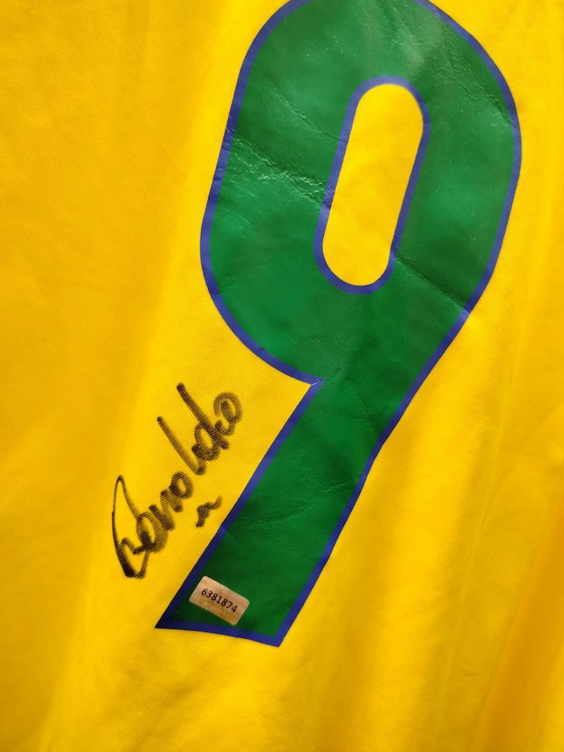 K&ouml;nnte beinhalten: Gelbes Trikot mit einer gro&szlig;en gr&uuml;nen Zahl 9, die blau umrandet ist. Das Trikot hat eine schwarze Signatur "Ronaldo". Ein kleiner Aufkleber mit der Nummer "6381874" ist ebenfalls sichtbar.