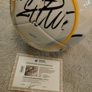 Puede incluir: Un bal&oacute;n de f&uacute;tbol blanco y gris con firma negra y detalles dorados. Se incluye un certificado de autenticidad, con el texto "GCOA Certificate of Authenticity" y el n&uacute;mero de serie de certificaci&oacute;n 6381818.