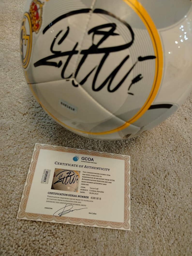 Puede incluir: Un bal&oacute;n de f&uacute;tbol blanco y gris con firma negra y detalles dorados. Se incluye un certificado de autenticidad, con el texto "GCOA Certificate of Authenticity" y el n&uacute;mero de serie de certificaci&oacute;n 6381818.