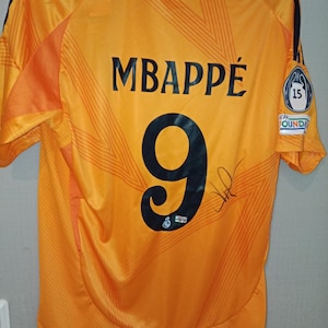 Pode incluir: Camisola de futebol laranja com o nome "MBAPPÉ" e o número "9" impressos em preto nas costas. A camisola tem uma assinatura e um patch com o número "15". Listras azuis escuras nos ombros.