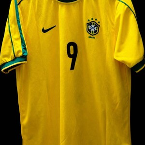 K&ouml;nnte beinhalten: Ein gelbes Brasilien-Fu&szlig;balltrikot mit gr&uuml;nen Verzierungen an Kragen und &Auml;rmeln. Die Nummer 9 ist schwarz auf der Vorderseite aufgedruckt. Ein schwarzer Nike-Swoosh befindet sich auf der rechten Brust, und ein brasilianisches Teamwappen auf der linken Brust.