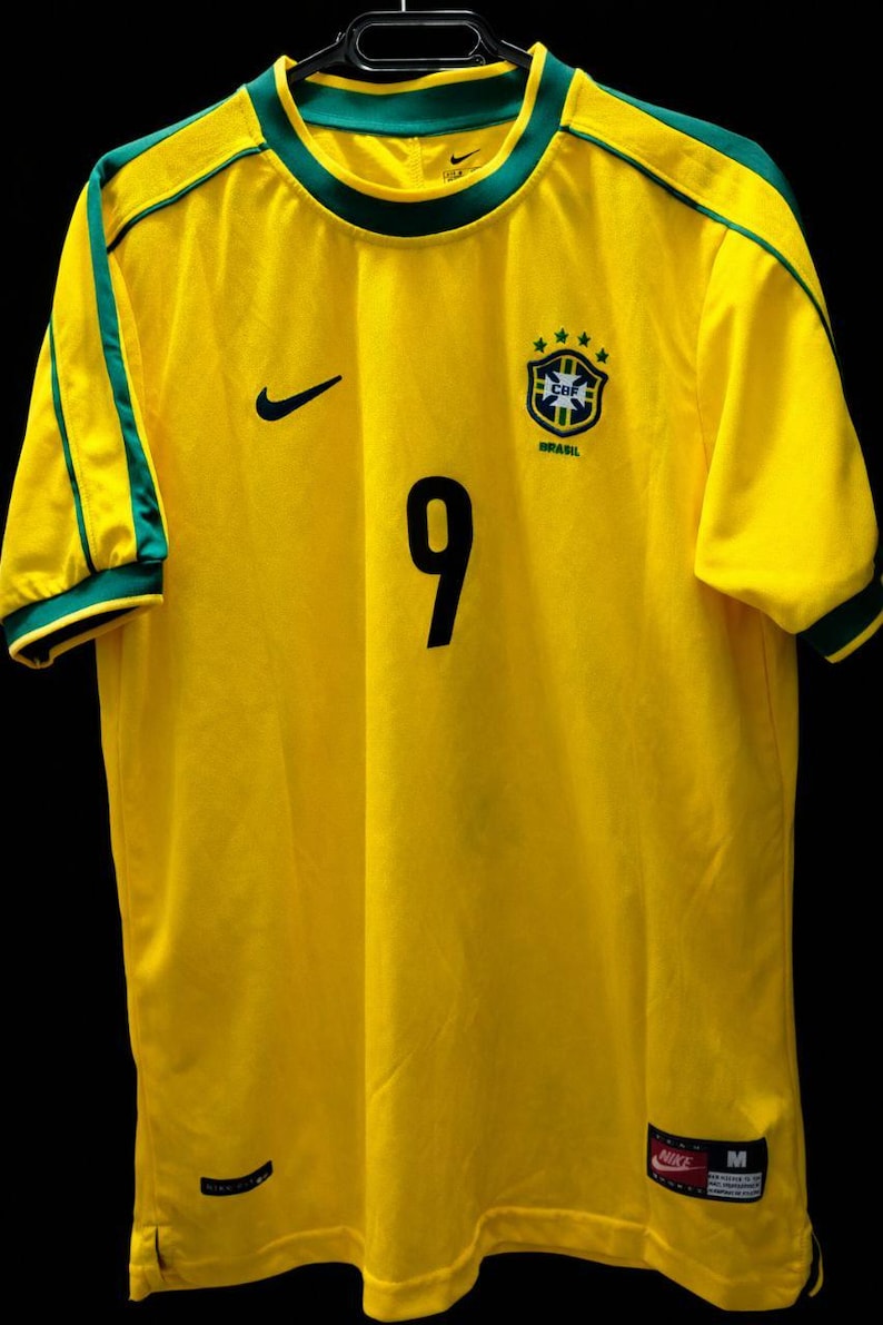 K&ouml;nnte beinhalten: Ein gelbes Brasilien-Fu&szlig;balltrikot mit gr&uuml;nen Verzierungen an Kragen und &Auml;rmeln. Die Nummer 9 ist schwarz auf der Vorderseite aufgedruckt. Ein schwarzer Nike-Swoosh befindet sich auf der rechten Brust, und ein brasilianisches Teamwappen auf der linken Brust.