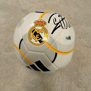 Puede incluir: Bal&oacute;n de f&uacute;tbol blanco y gris con el logo del Real Madrid, una corona dorada y detalles en negro. El bal&oacute;n tiene una firma negra y rayas amarillas. El bal&oacute;n est&aacute; sobre una superficie marr&oacute;n claro.