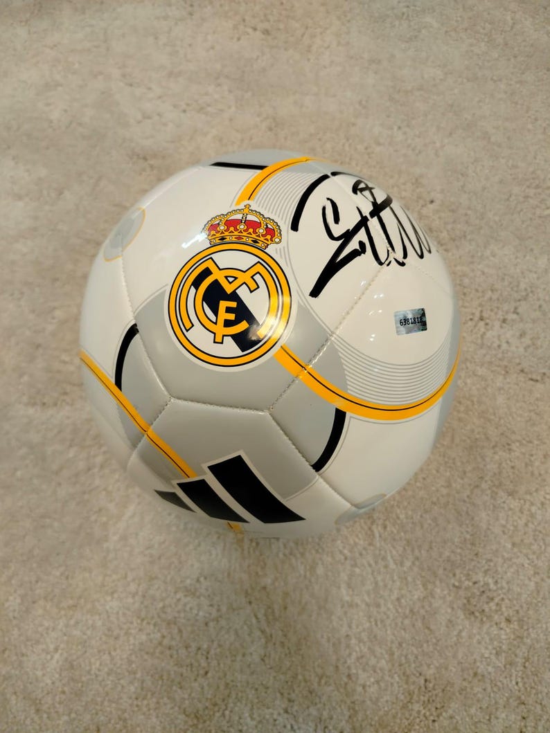 Puede incluir: Bal&oacute;n de f&uacute;tbol blanco y gris con el logo del Real Madrid, una corona dorada y detalles en negro. El bal&oacute;n tiene una firma negra y rayas amarillas. El bal&oacute;n est&aacute; sobre una superficie marr&oacute;n claro.