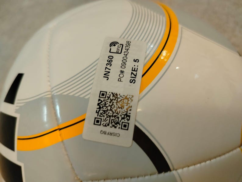 Puede incluir: Un bal&oacute;n de f&uacute;tbol blanco y gris con detalles en negro y amarillo. Una etiqueta adjunta con el texto "JN7360", "TALLA: 5" y un c&oacute;digo QR. El bal&oacute;n est&aacute; dise&ntilde;ado para deportes y recreaci&oacute;n.