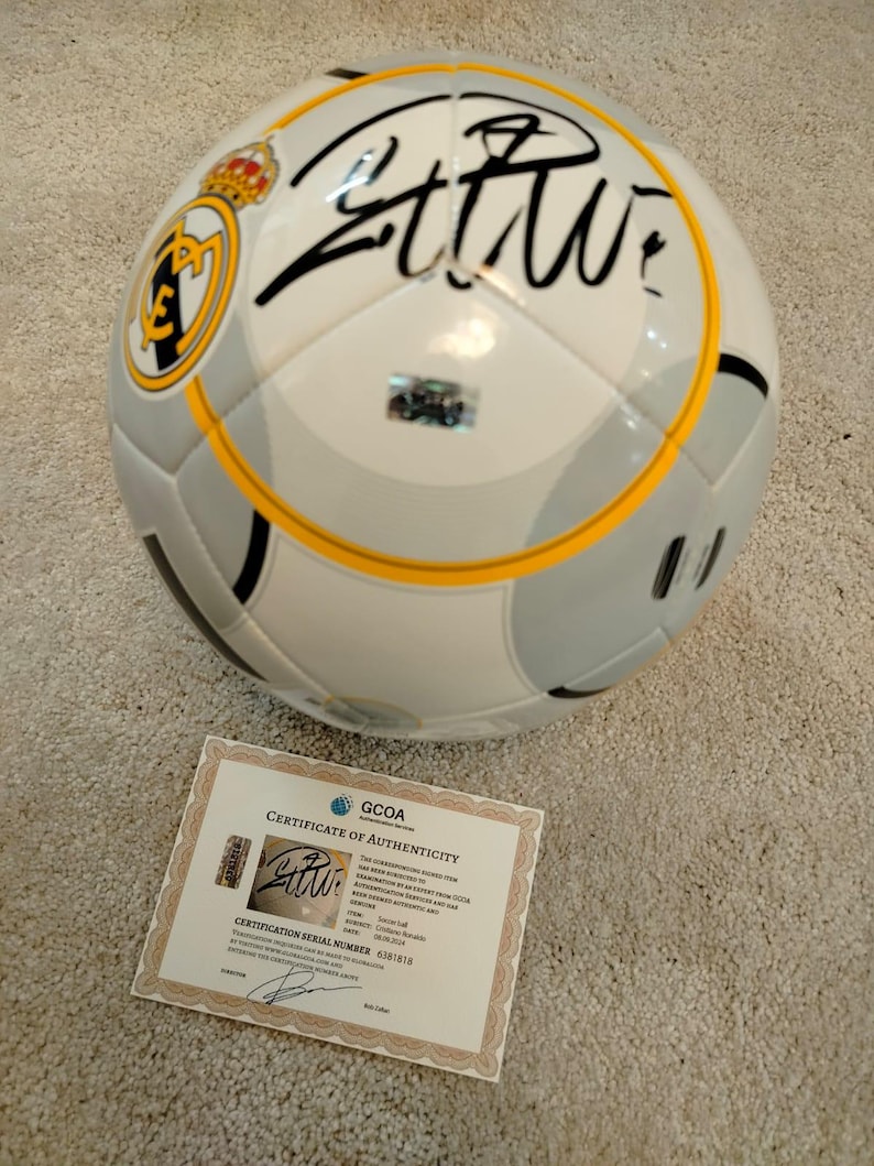 Puede incluir: Bal&oacute;n de f&uacute;tbol blanco y gris con el logo del Real Madrid y una firma negra. El bal&oacute;n tiene una franja amarilla. Se incluye un certificado de autenticidad.