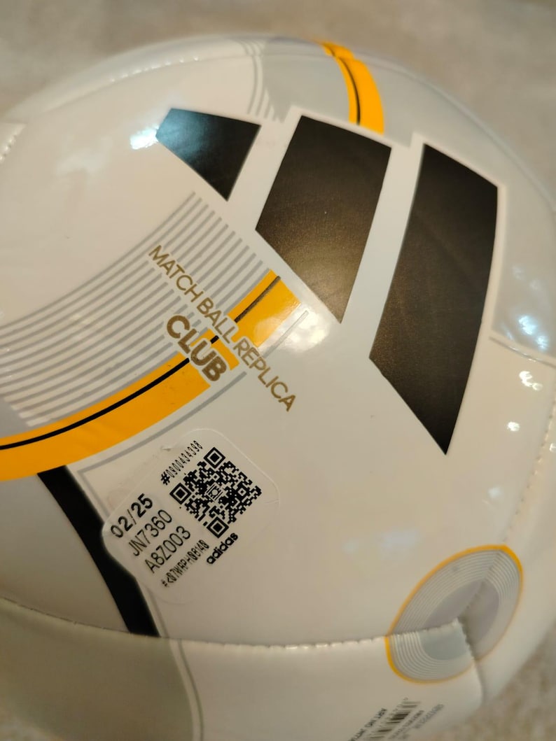 Puede incluir: Bal&oacute;n de f&uacute;tbol blanco y gris con el logotipo negro de Adidas y detalles amarillos. El bal&oacute;n tiene el texto "MATCH BALL REPLICA CLUB" y una pegatina con c&oacute;digo QR. Dise&ntilde;ado para el juego de clubes.