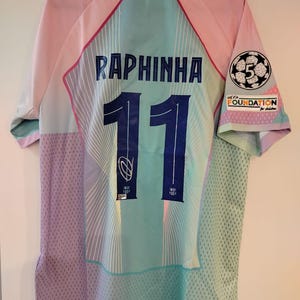 May include: A pastel-colored soccer jersey with the name "RAPHINHA" and the number "11" on the back. The jersey features a light blue, pink, and purple color scheme with a "UEFA FOUNDATION for children" patch on the sleeve. The jersey is hanging on a black hanger.