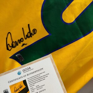 K&ouml;nnte beinhalten: Ein gelbes Fu&szlig;balltrikot mit einer gro&szlig;en gr&uuml;nen Nummer 9 und blauem Umriss. Das Trikot hat eine Unterschrift und ein Echtheitszertifikat von GCOA. Das Zertifikat best&auml;tigt, dass es sich um ein Ronaldo Nazario Trikot handelt.