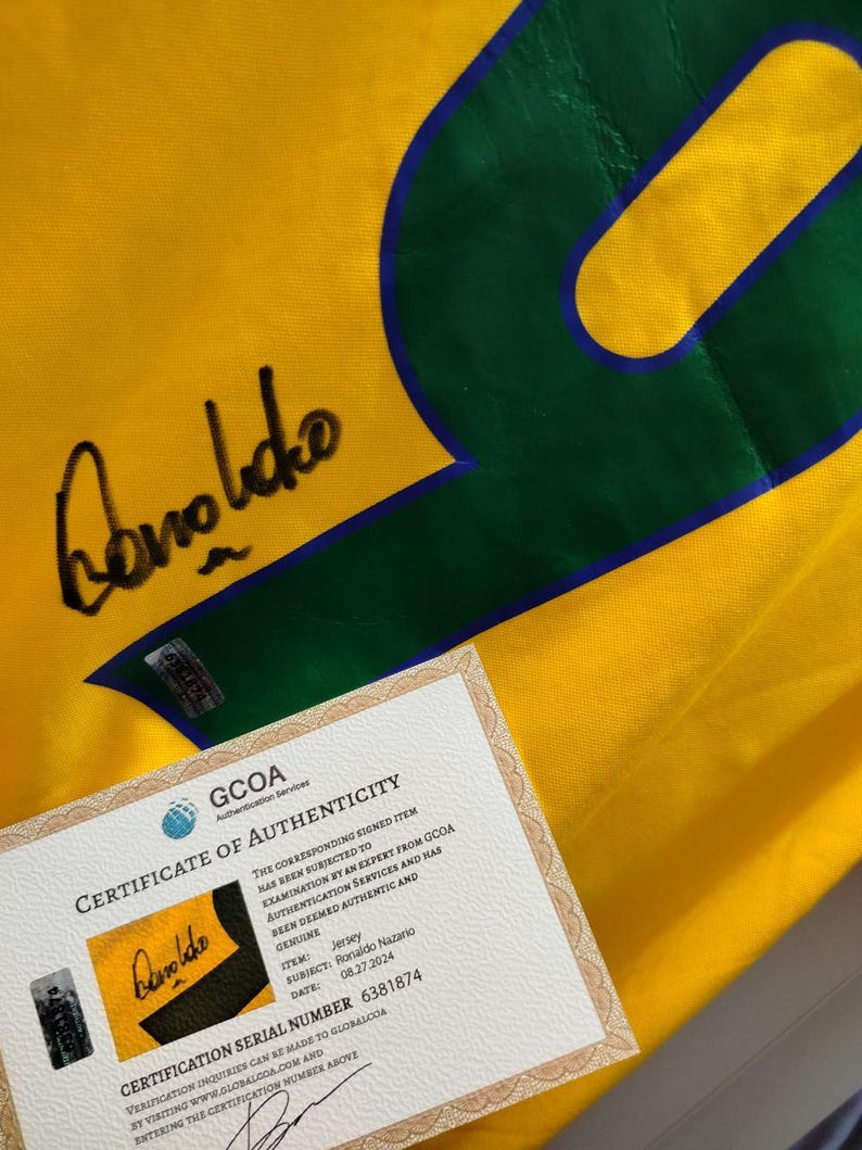 K&ouml;nnte beinhalten: Ein gelbes Fu&szlig;balltrikot mit einer gro&szlig;en gr&uuml;nen Nummer 9 und blauem Umriss. Das Trikot hat eine Unterschrift und ein Echtheitszertifikat von GCOA. Das Zertifikat best&auml;tigt, dass es sich um ein Ronaldo Nazario Trikot handelt.