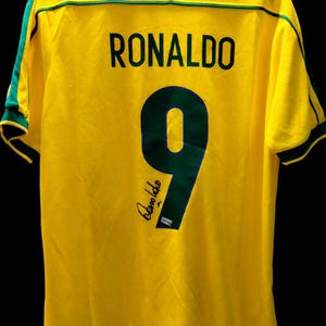 K&ouml;nnte beinhalten: Ein gelbes Fu&szlig;balltrikot mit gr&uuml;nen Verzierungen, das den Namen "RONALDO" und die Nummer "9" in Dunkelgr&uuml;n zeigt. Das Trikot hat auch eine Unterschrift in der unteren linken Ecke. Das Trikot h&auml;ngt vor einem schwarzen Hintergrund.