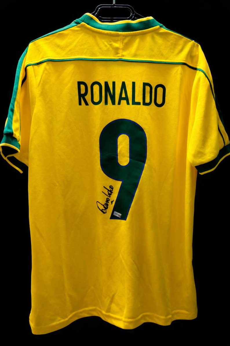 K&ouml;nnte beinhalten: Ein gelbes Fu&szlig;balltrikot mit gr&uuml;nen Verzierungen, das den Namen "RONALDO" und die Nummer "9" in Dunkelgr&uuml;n zeigt. Das Trikot hat auch eine Unterschrift in der unteren linken Ecke. Das Trikot h&auml;ngt vor einem schwarzen Hintergrund.