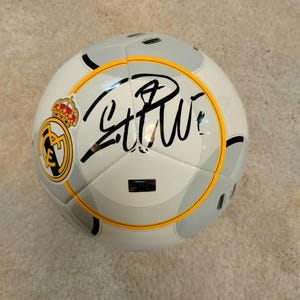 Puede incluir: Un bal&oacute;n de f&uacute;tbol blanco y gris con detalles en negro y un emblema amarillo y negro. El bal&oacute;n presenta una firma en tinta negra y una corona dorada. El bal&oacute;n est&aacute; dise&ntilde;ado para f&uacute;tbol.