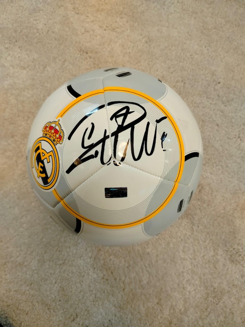 Puede incluir: Un bal&oacute;n de f&uacute;tbol blanco y gris con detalles en negro y un emblema amarillo y negro. El bal&oacute;n presenta una firma en tinta negra y una corona dorada. El bal&oacute;n est&aacute; dise&ntilde;ado para f&uacute;tbol.